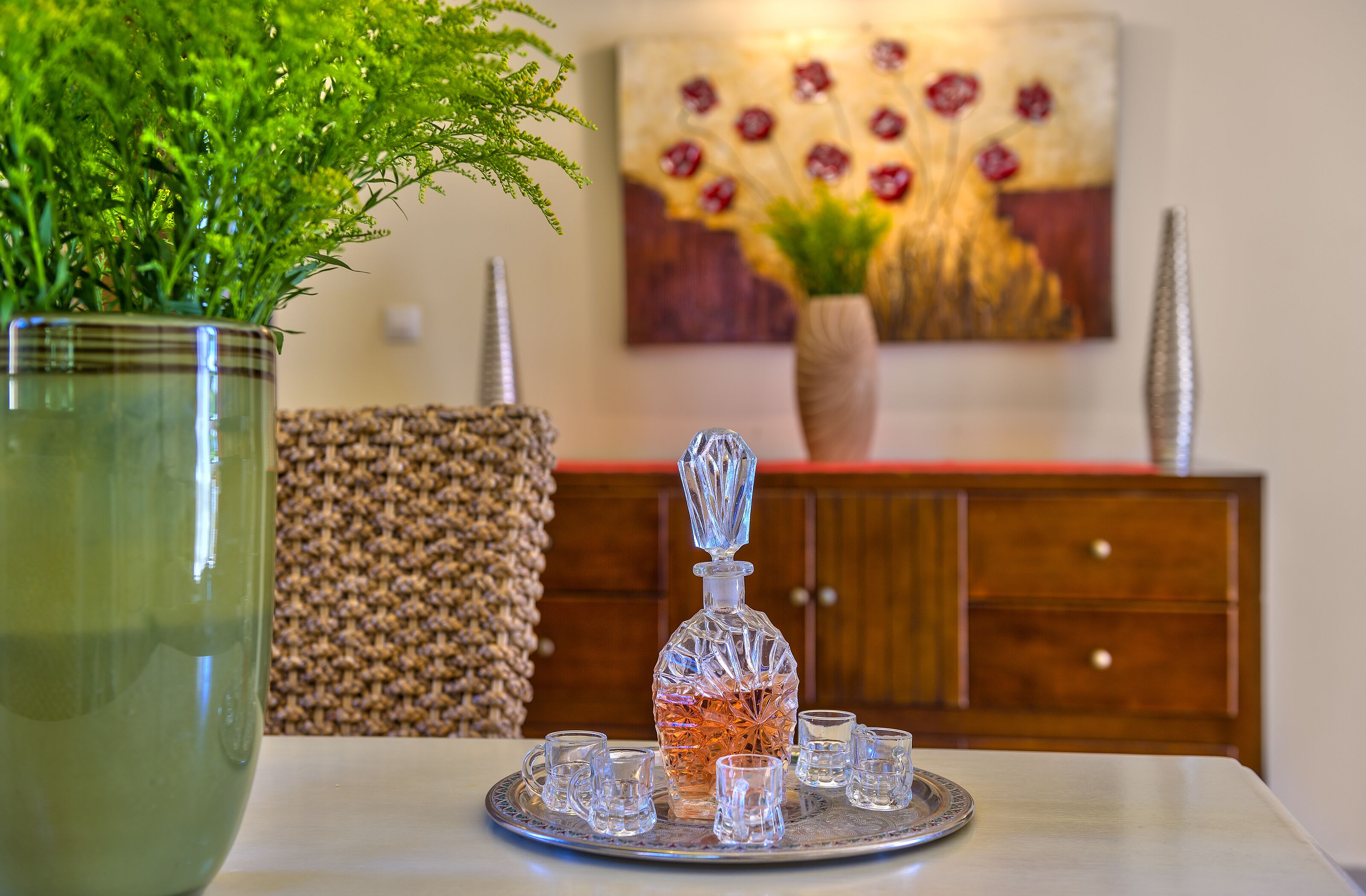 A dining table with a glass decanter, six glasses, and a large green plant.
