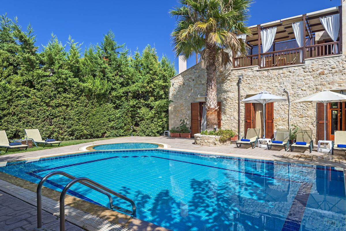 Une piscine entourée de chaises longues et de parasols, bordée d’un bâtiment en pierre avec des volets en bois et un balcon agrémenté de rideaux blancs.