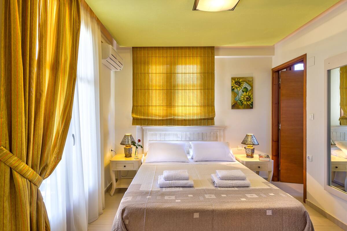 Une chambre d’hôtel avec un lit, deux tables de chevet avec des lampes, une grande fenêtre avec des rideaux, un tableau au mur et une porte en bois.