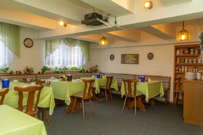 Uma área de refeições com mesas para quatro pessoas, cadeiras de madeira, toalhas de mesa verdes, um projetor no teto e uma estante com utensílios de cozinha.