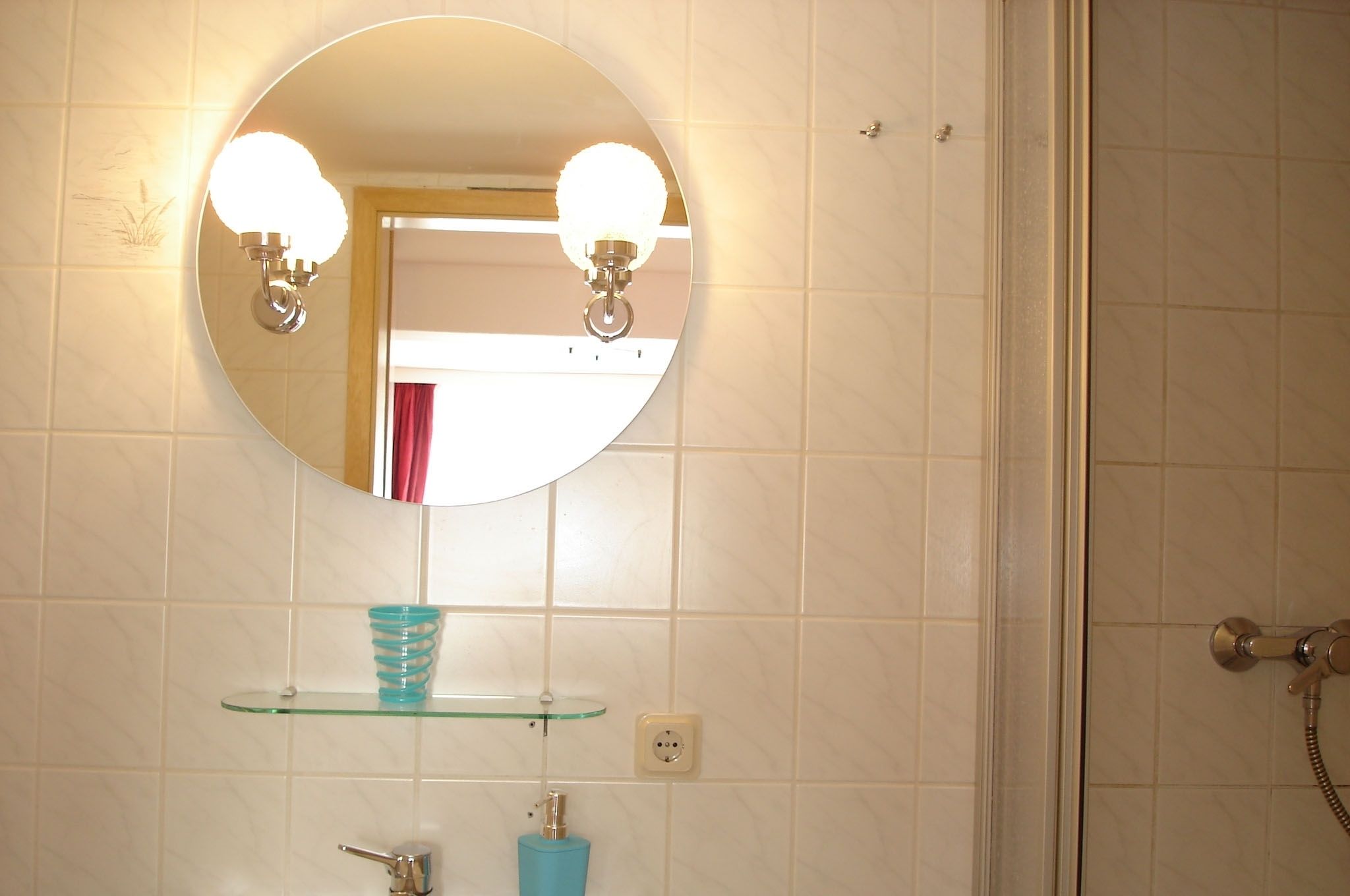 A bathroom with a round mirror, two wall-mounted lights, a glass shelf, a faucet, and a shower control.