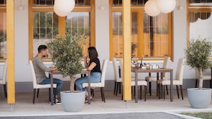 Un café al aire libre con mesas y sillas de madera, plantas en macetas y lámparas colgantes.