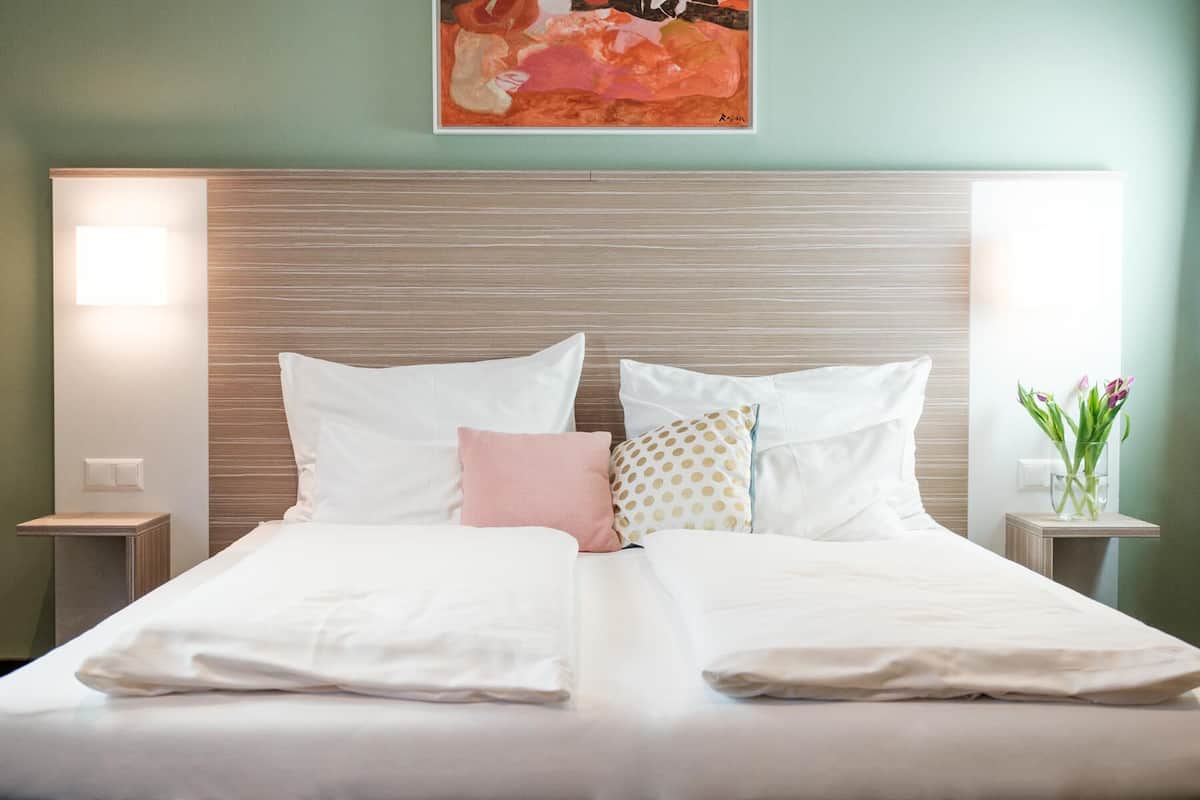 A neatly made double bed with white linens, a wooden headboard, and a pink pillow. There are two bedside tables with lamps, and a vase with flowers on the right side.