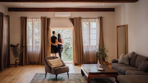 Un salón amplio con suelo de madera, un sofá, una mesa de centro y un sillón. Hay una ventana grande con cortinas, una planta en maceta y un espejo en la pared.
