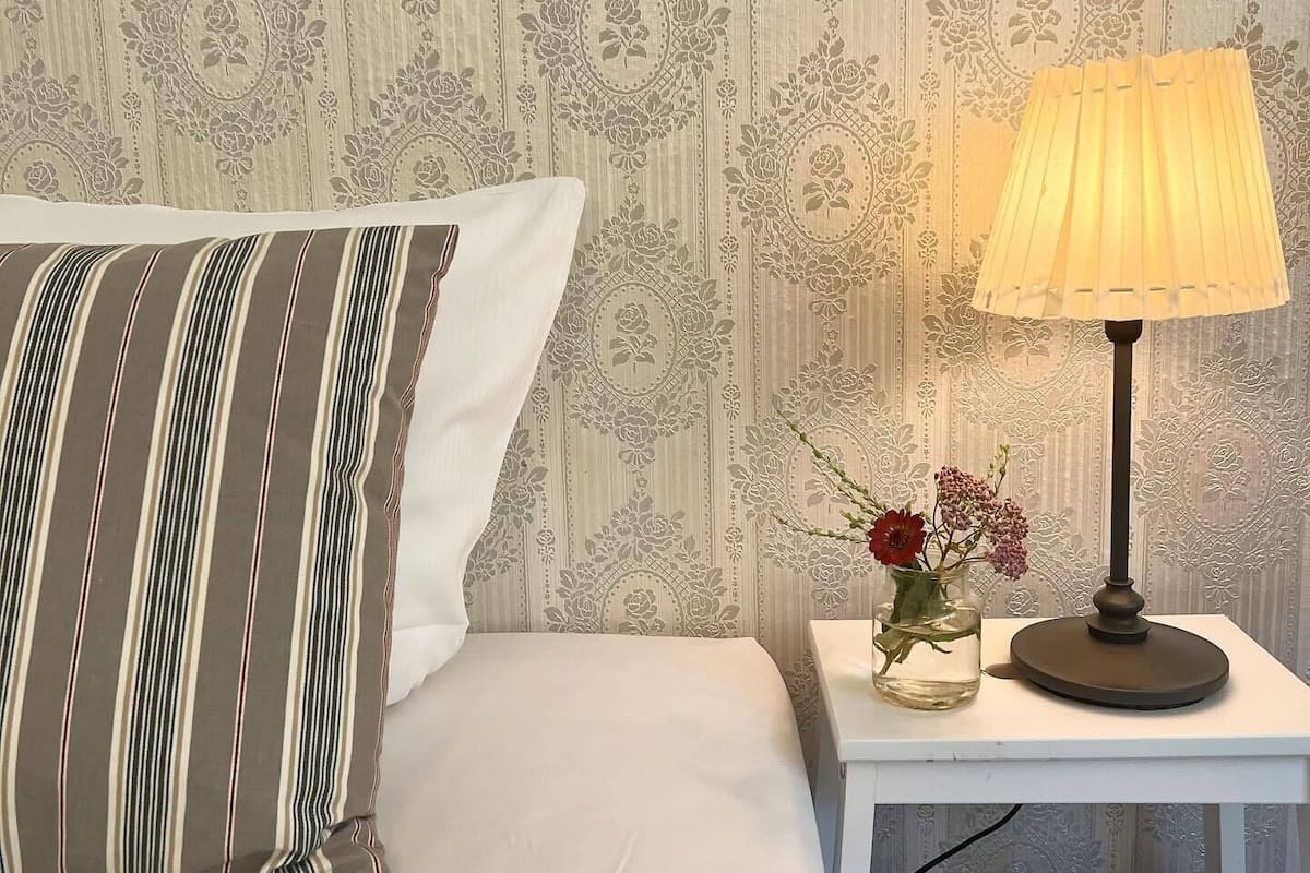 A neatly made bed with striped pillows, a bedside table with a lamp and a vase of flowers, and a wallpapered wall with a floral pattern.