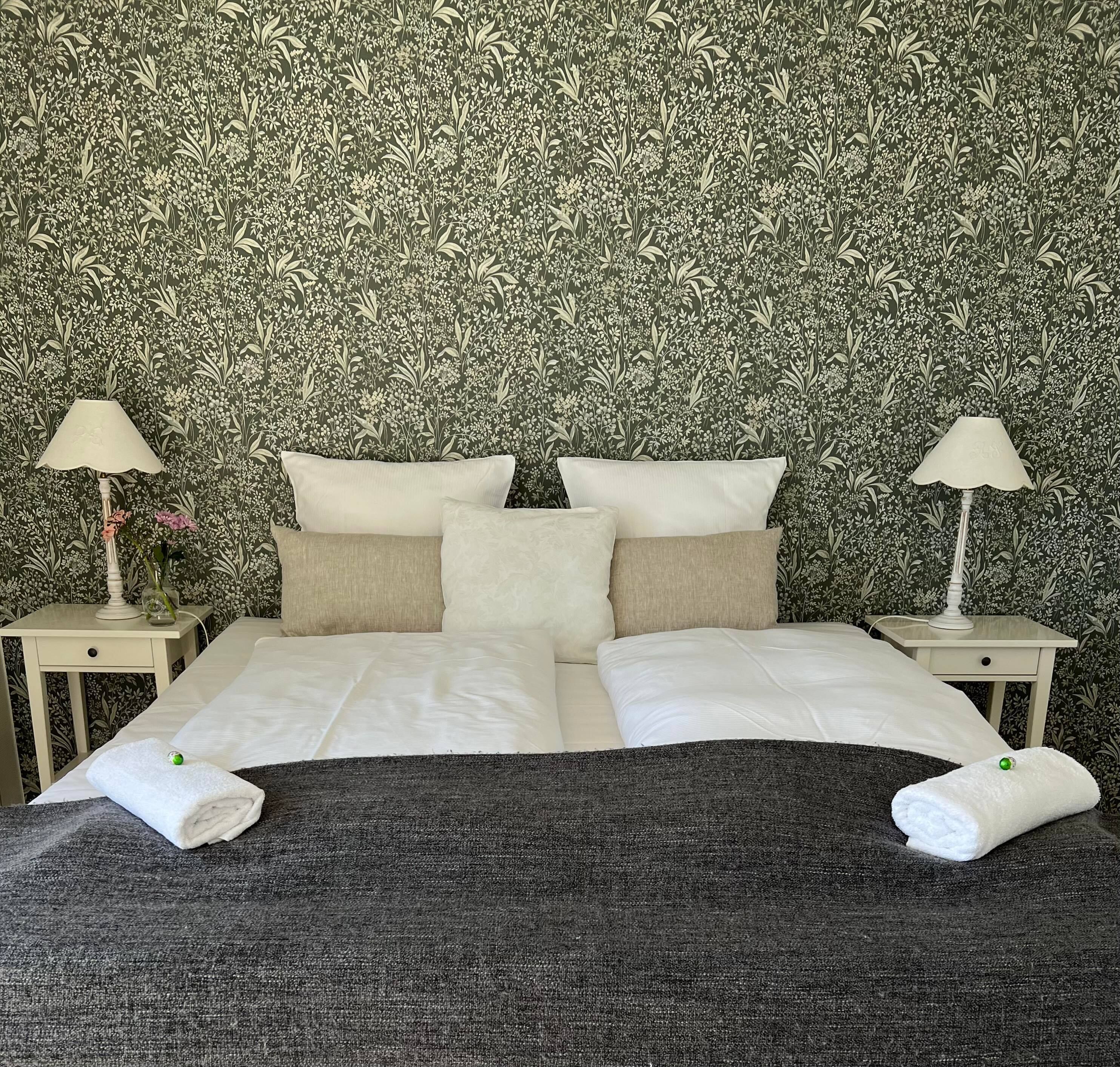 A neatly made bed with white linens, two bedside tables with lamps, and a floral wallpaper background.