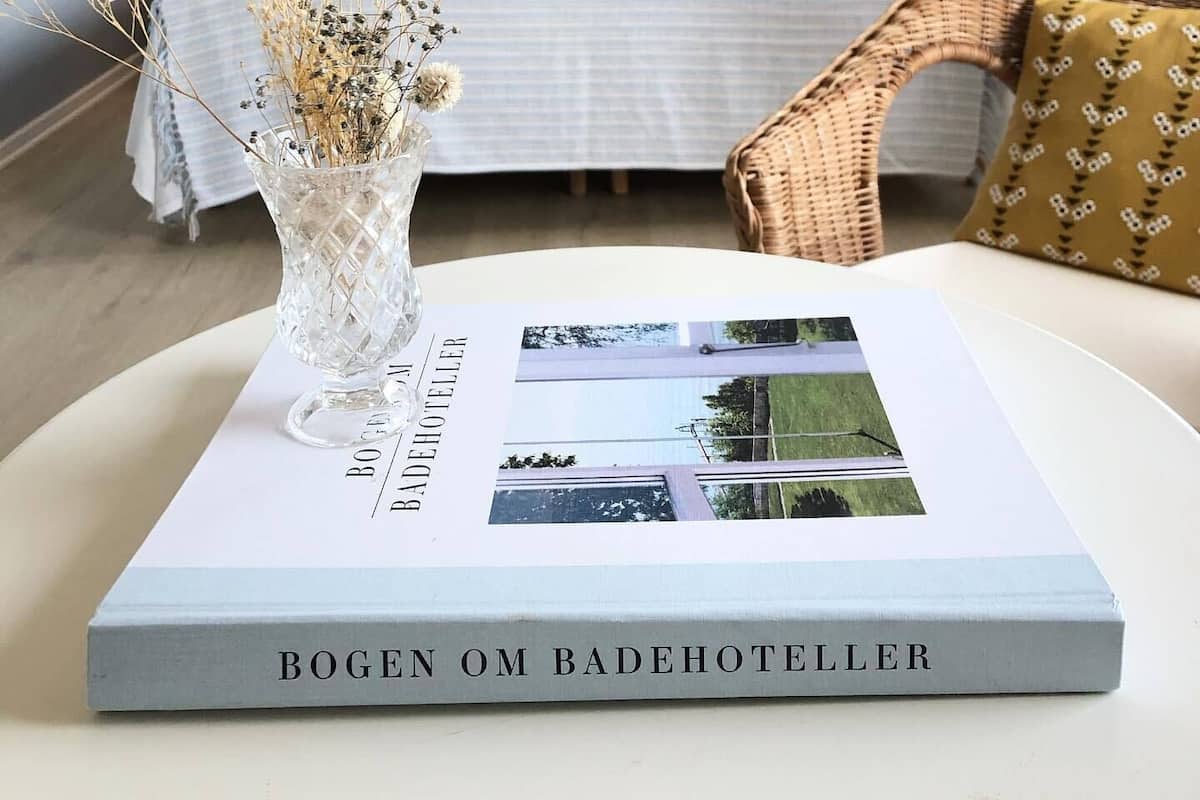 A hotel room with a bed, a wicker chair, a vase with dried flowers, and a book titled "BOGEN OM BADEHOTELLER".