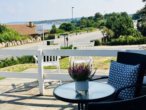 A patio with white chairs and a table, overlooking a road and greenery. - Sandkaas Badehotel (Allinge)