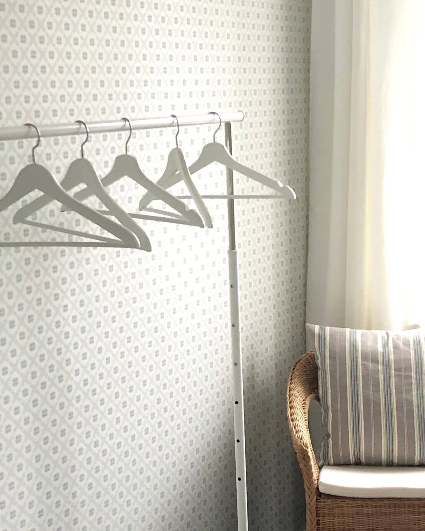 A room with a patterned wallpaper, a white metal drying rack with hangers, and a wicker chair with a striped cushion.