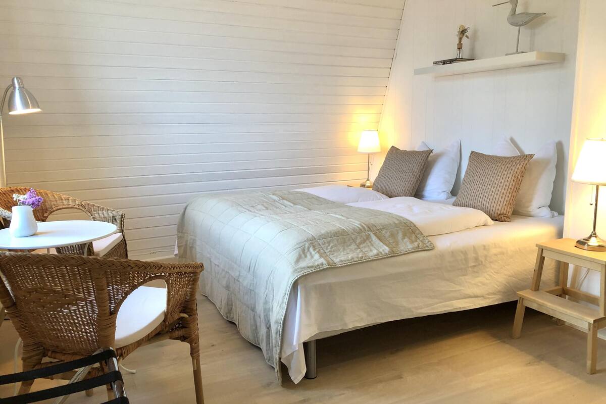A neatly arranged bedroom with a bed, a small table with a vase, wicker chairs, and a nightstand with a lamp.