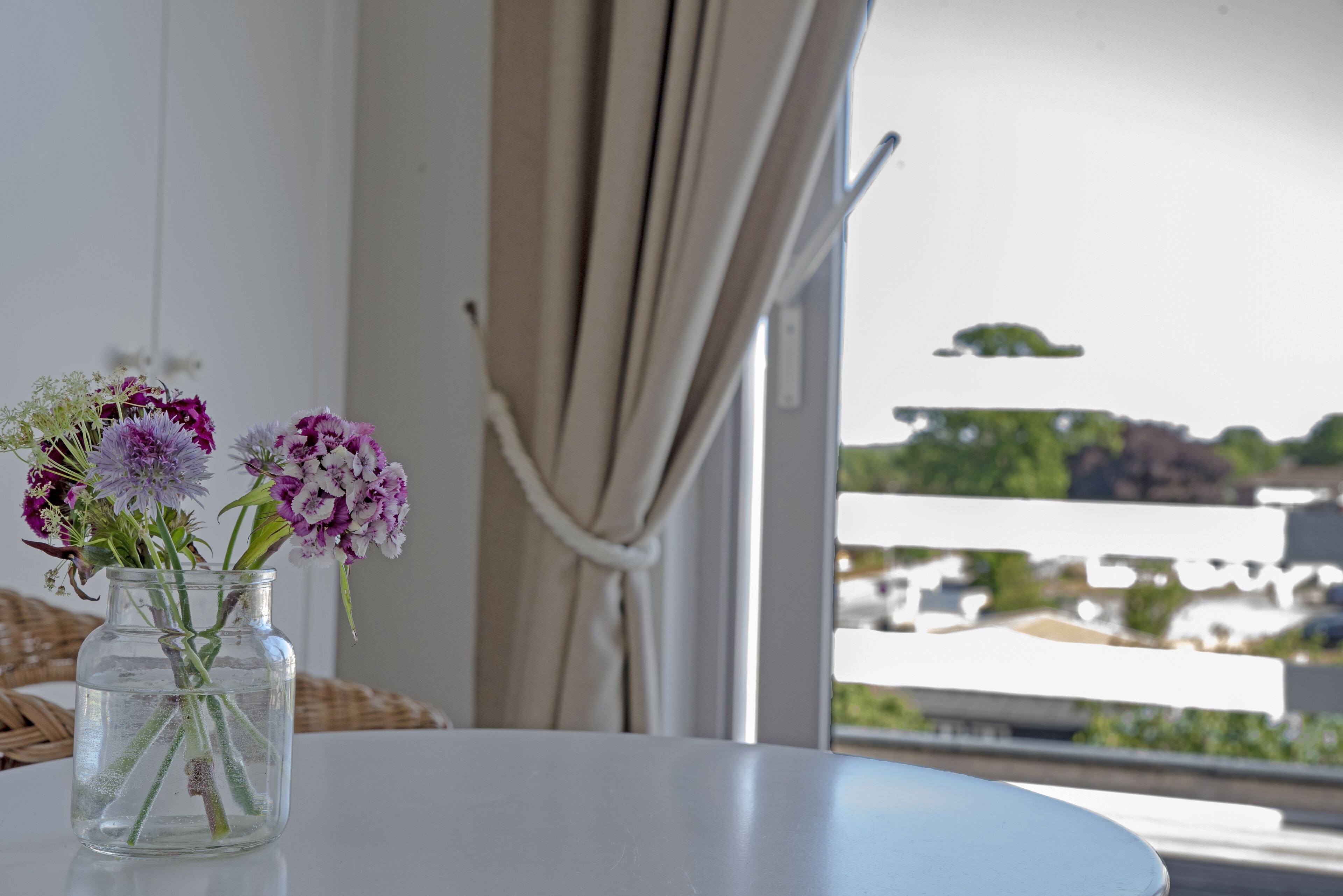 On voit clairement un balcon avec une balustrade blanche, une chaise en osier et un vase rempli de fleurs posé sur une table.