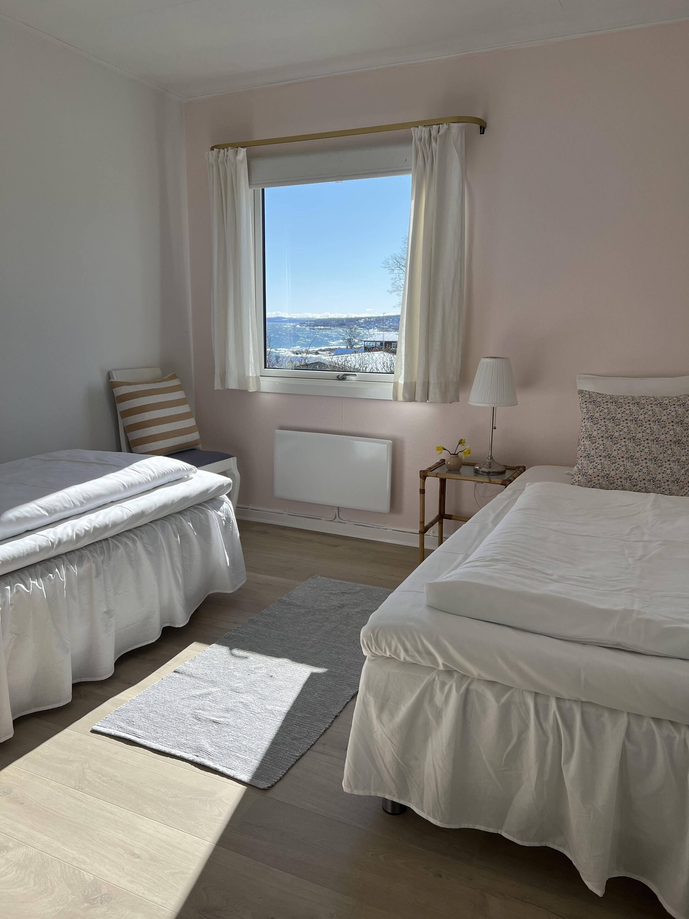 A bedroom with two beds, a window with curtains, a radiator, a lamp, and a view of the outside.