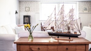 Une table en bois sur laquelle est posé un modèle de bateau, ainsi qu’un vase rempli de fleurs jaunes, dans un salon où se trouvent un canapé blanc et un papier peint à motifs.
