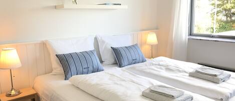 Una cama doble con ropa de cama blanca y dos almohadas, una mesita de noche con una lámpara, y una ventana con vistas a árboles.