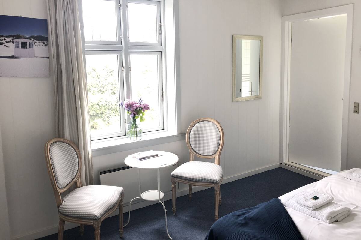 A hotel room with a bed, two chairs, a small table, a window with curtains, a mirror, and a radiator.