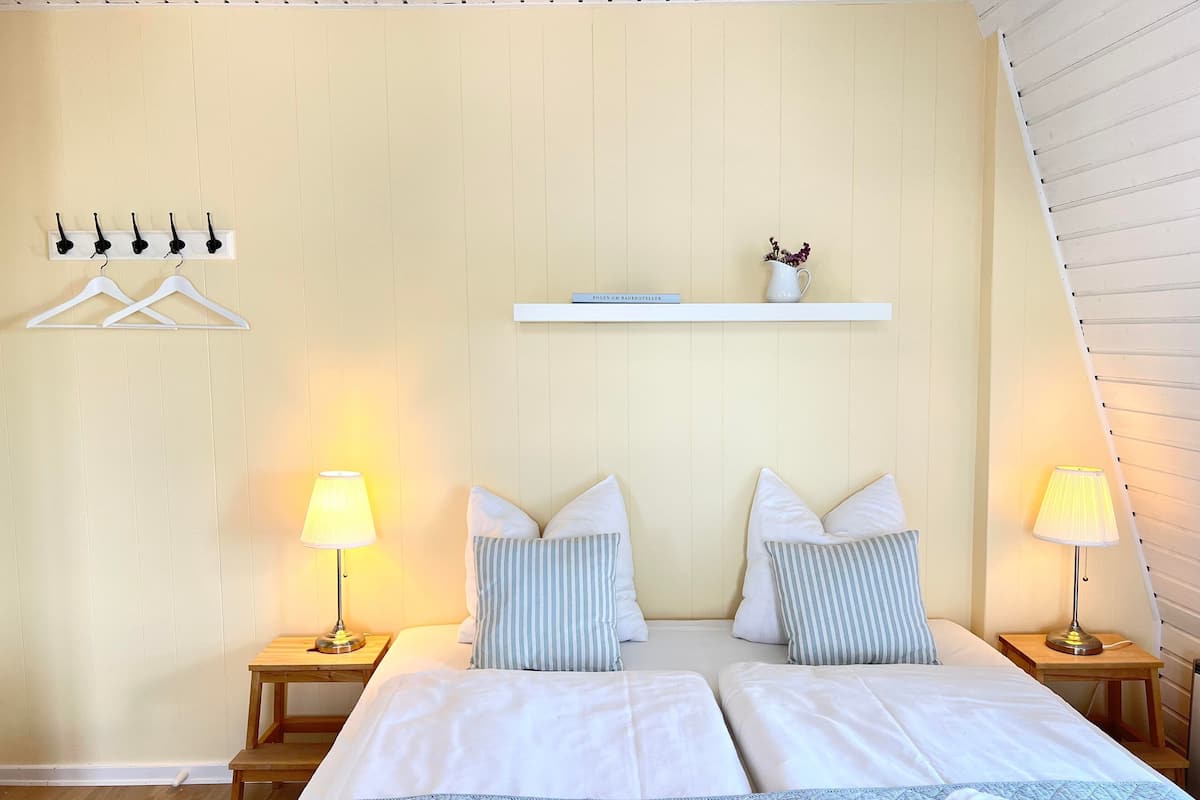 A double bed with two pillows and a bedside table with a lamp. There is a shelf above the bed with a book and a small vase.