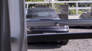 Une voiture garée dans une allée menant à une maison, avec vue sur l’océan et un parasol blanc.