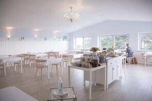 A bright, spacious room with white walls and wooden floors, featuring a kitchen area with various jars and a person working at the counter. - Sandkaas Badehotel (Allinge)