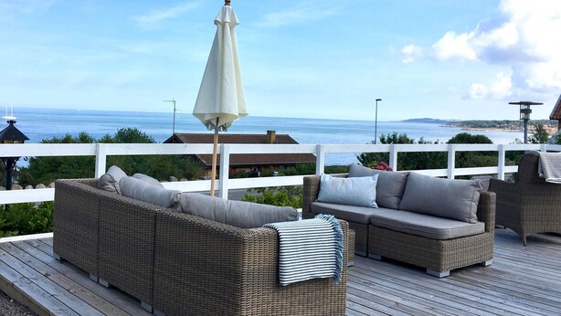Une terrasse dotée de meubles en osier et d’un parasol blanc, offrant une vue sur une étendue d’eau et une côte lointaine.