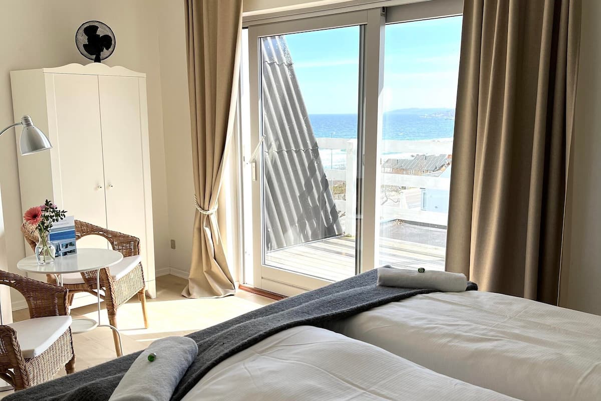 A hotel room with two beds, a desk with a lamp, a chair, a wardrobe, and a view of the sea.