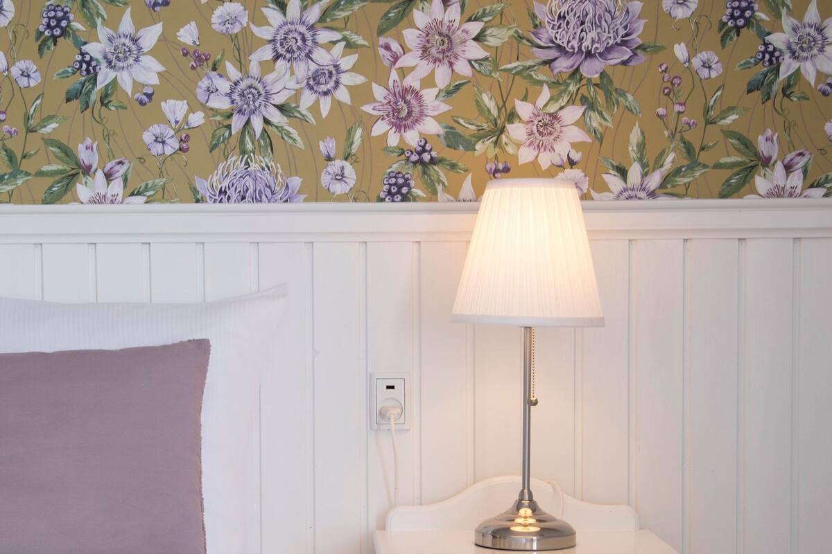 A neatly made bed with a white comforter and a pink pillow, a white nightstand with a lamp, and a floral wallpaper pattern.
