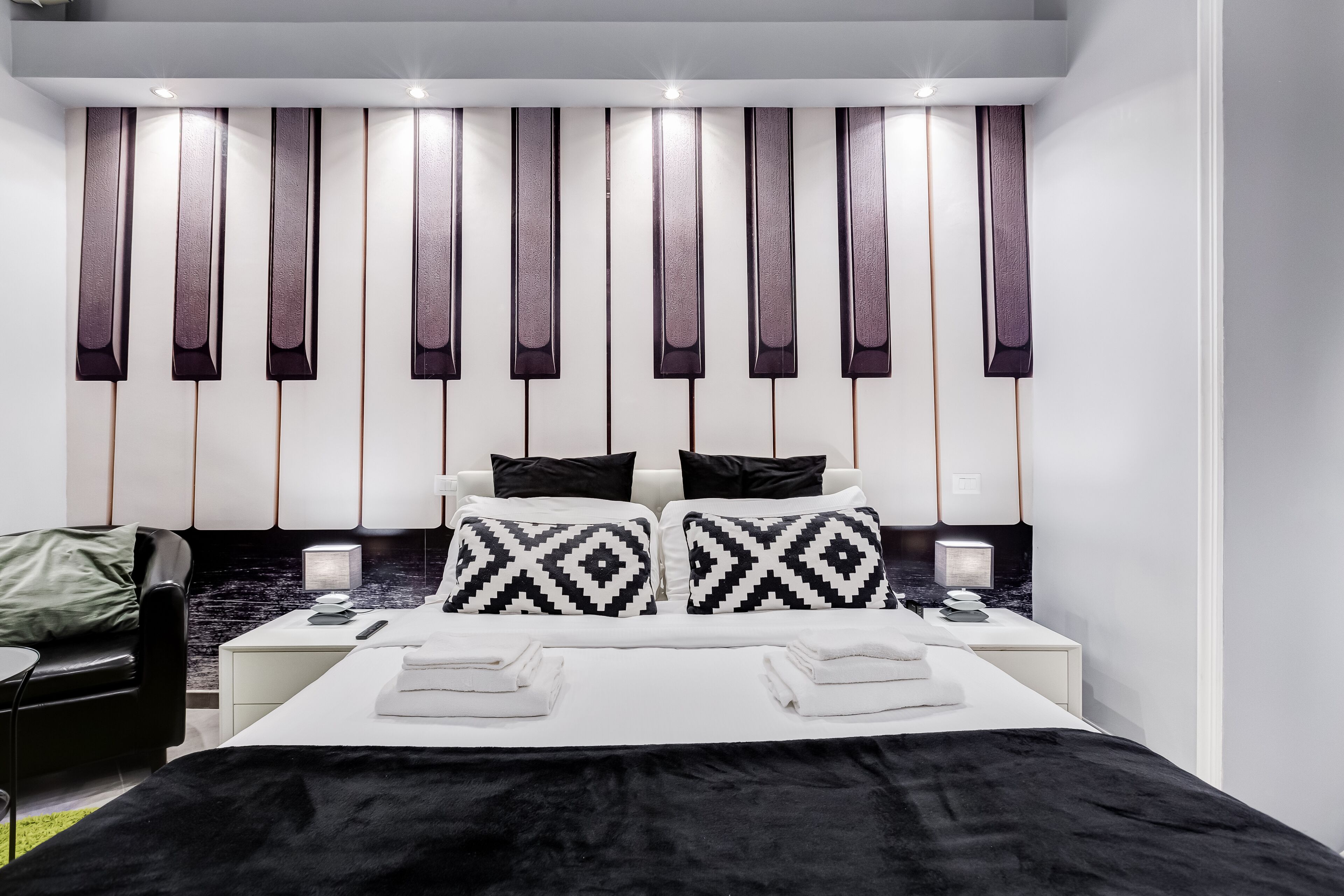 A modern hotel room with a large bed, two bedside tables, a black leather chair, and a wall with vertical light panels.