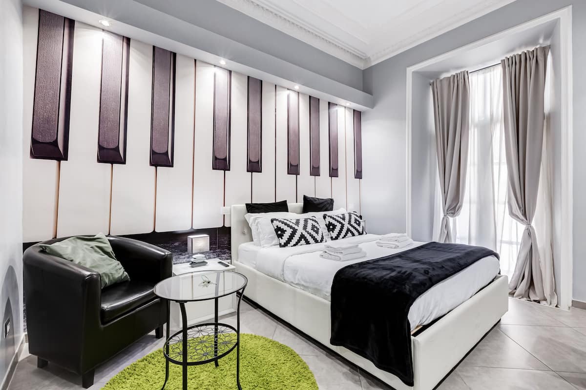 A modern bedroom with a white bed, black and white pillows, a black leather sofa, a small round table, and a green rug.