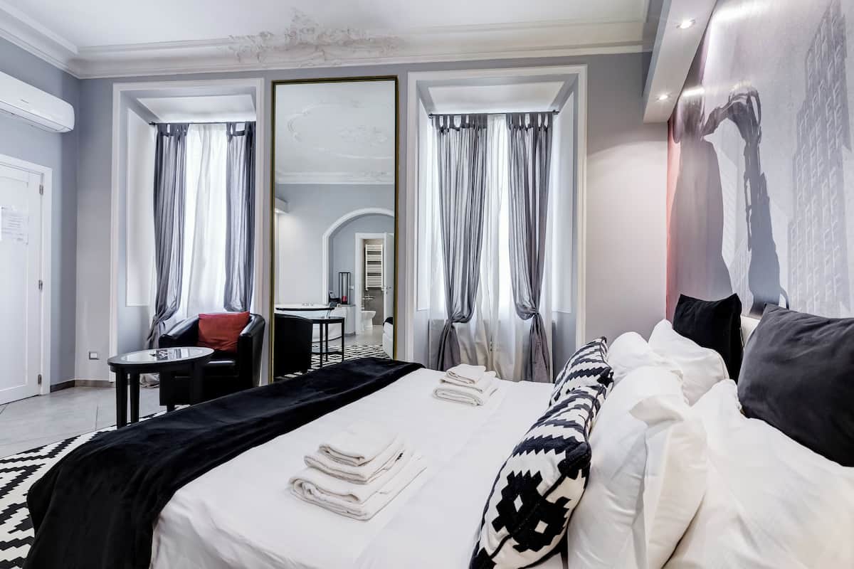 A modern hotel room with a large bed, a sitting area with a chair and table, and a mirror.
