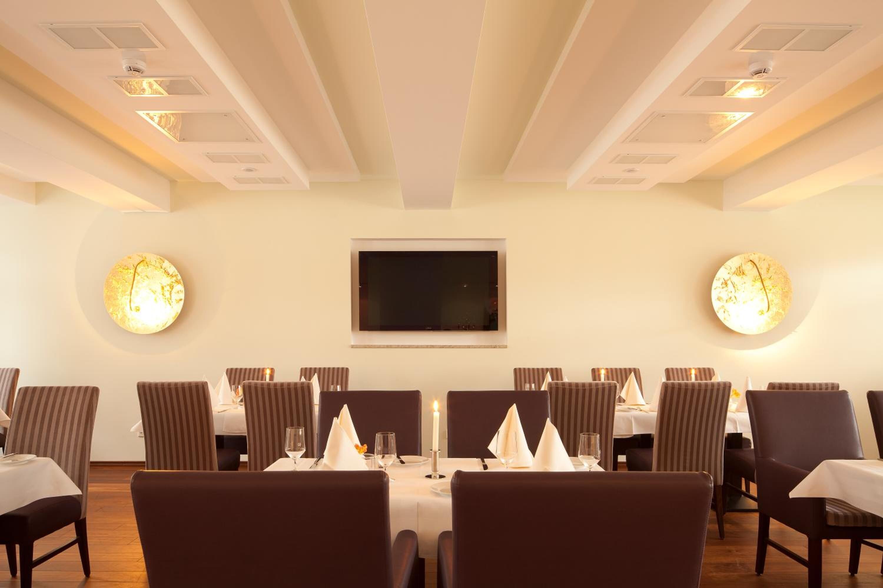 Un comedor con mesas dispuestas para seis personas, cada una con una vela, sillas y un televisor de pantalla plana montado en la pared.