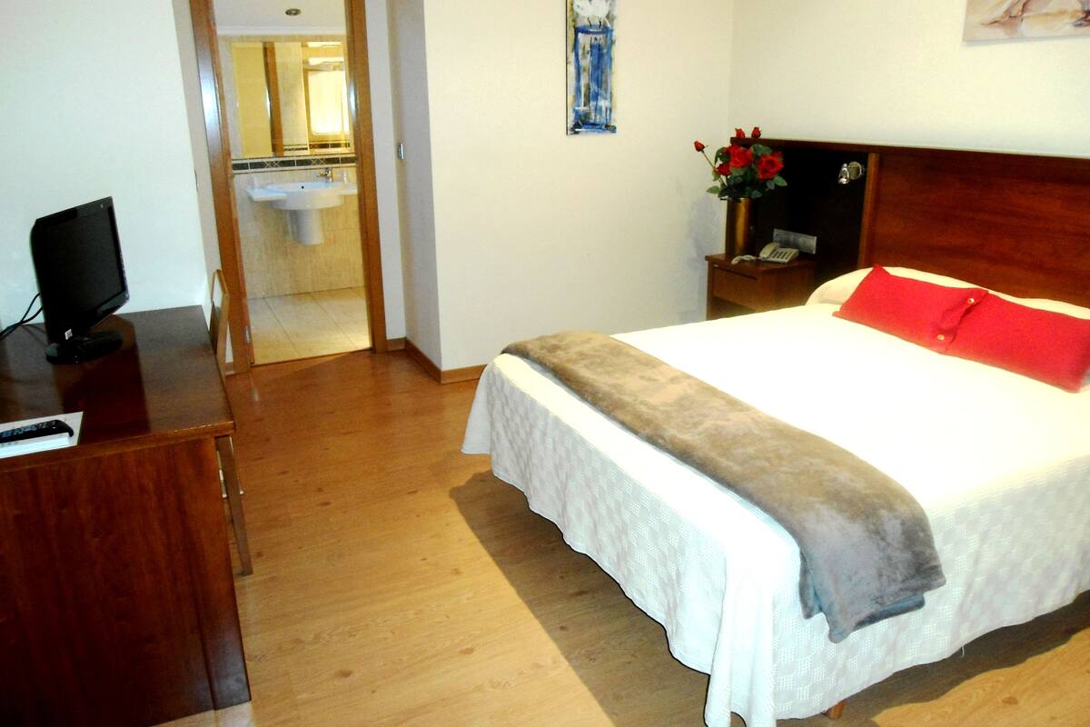 Una habitación de hotel con cama, escritorio con computadora, mesita de noche con un jarrón de flores, y un baño visible a través de una puerta abierta.