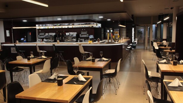Un restaurante con varias mesas y sillas, una zona de bar con mostrador y distintos elementos en exhibición, y un diseño interior moderno.