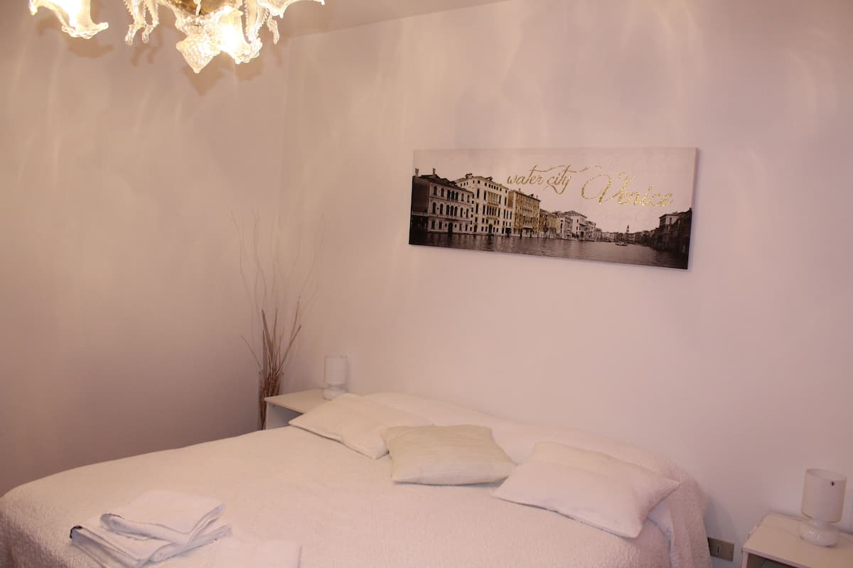 A bedroom with a bed, pillows, a wall-mounted picture of Venice, and a bedside table with a lamp.
