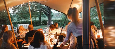 An outdoor dining area with tables under a canopy, surrounded by trees.
