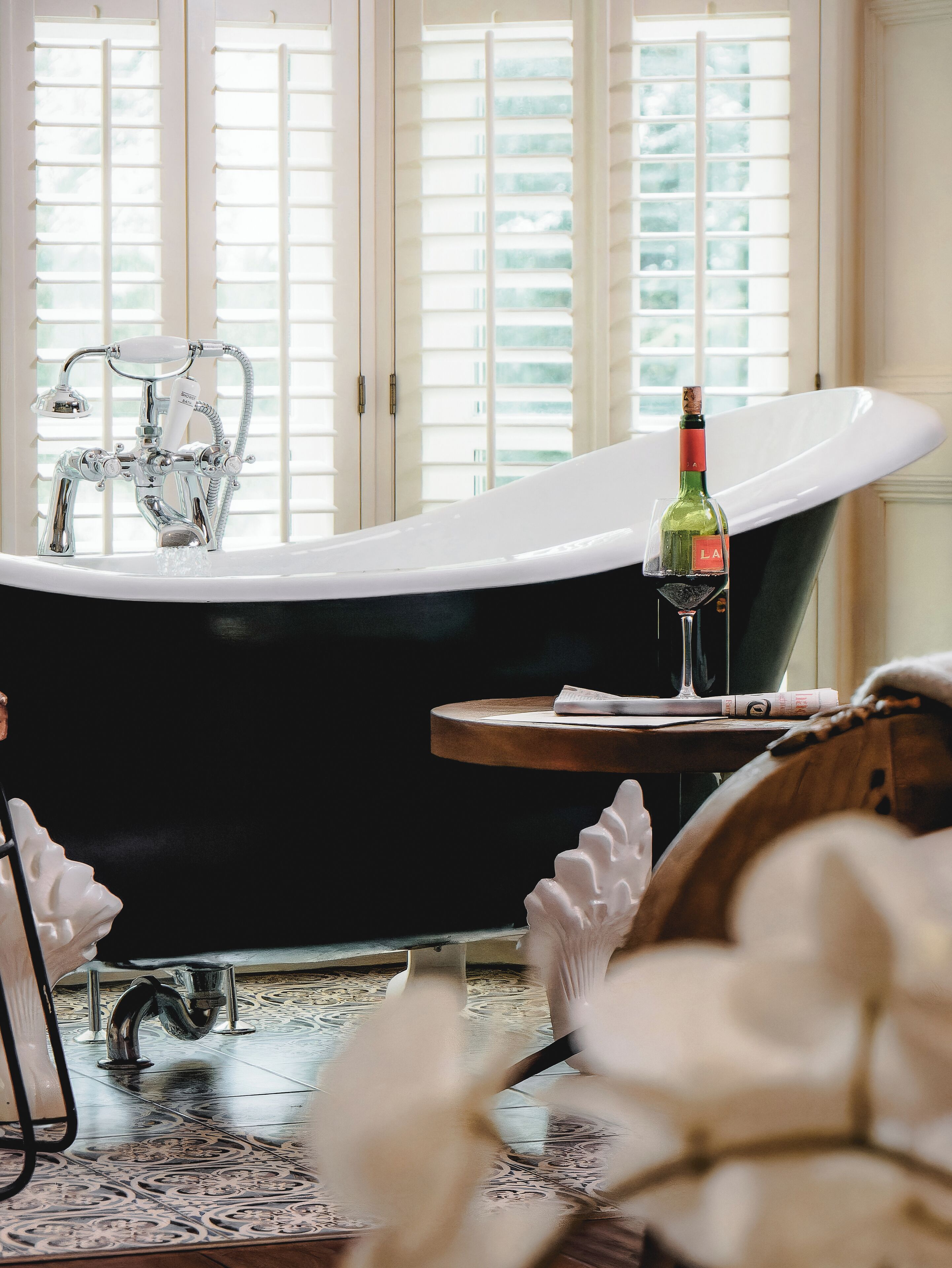 Une salle de bain avec une baignoire sur pieds, une bouteille de vin posée sur une petite table et une plante décorative.