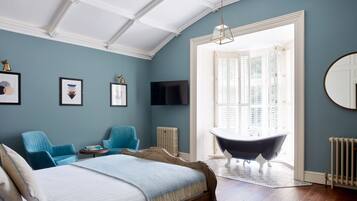 Un dormitorio con una cama grande, una bañera, dos sillas azules, un televisor y un ventanal con contraventanas.