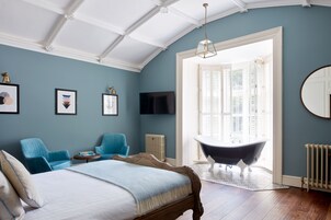Un dormitorio con una cama grande, una bañera, dos sillas azules, un televisor y un ventanal con contraventanas.