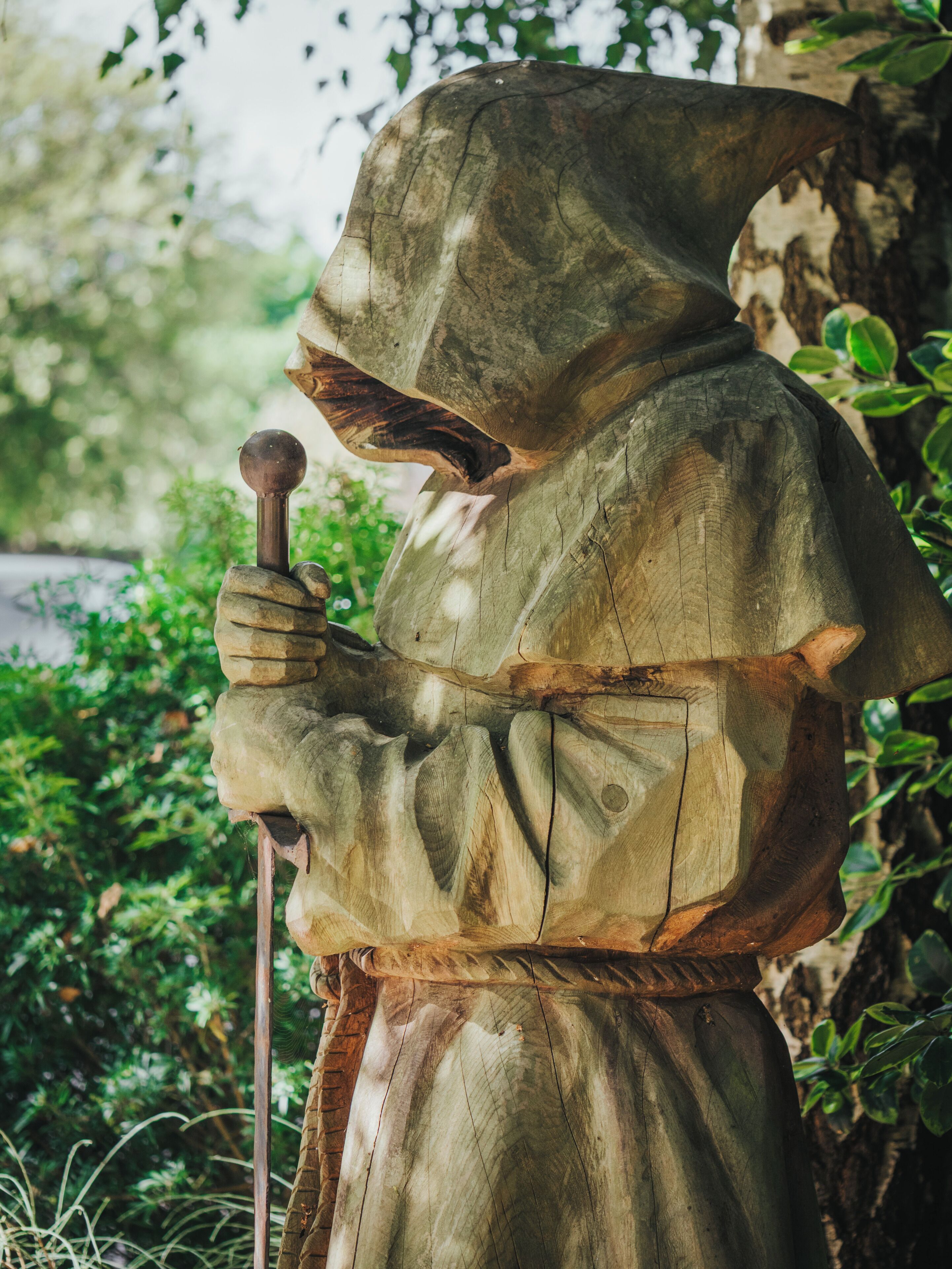 Une statue représentant une figure à capuche tenant un bâton, se dressant sur un fond de verdure et d’arbres.