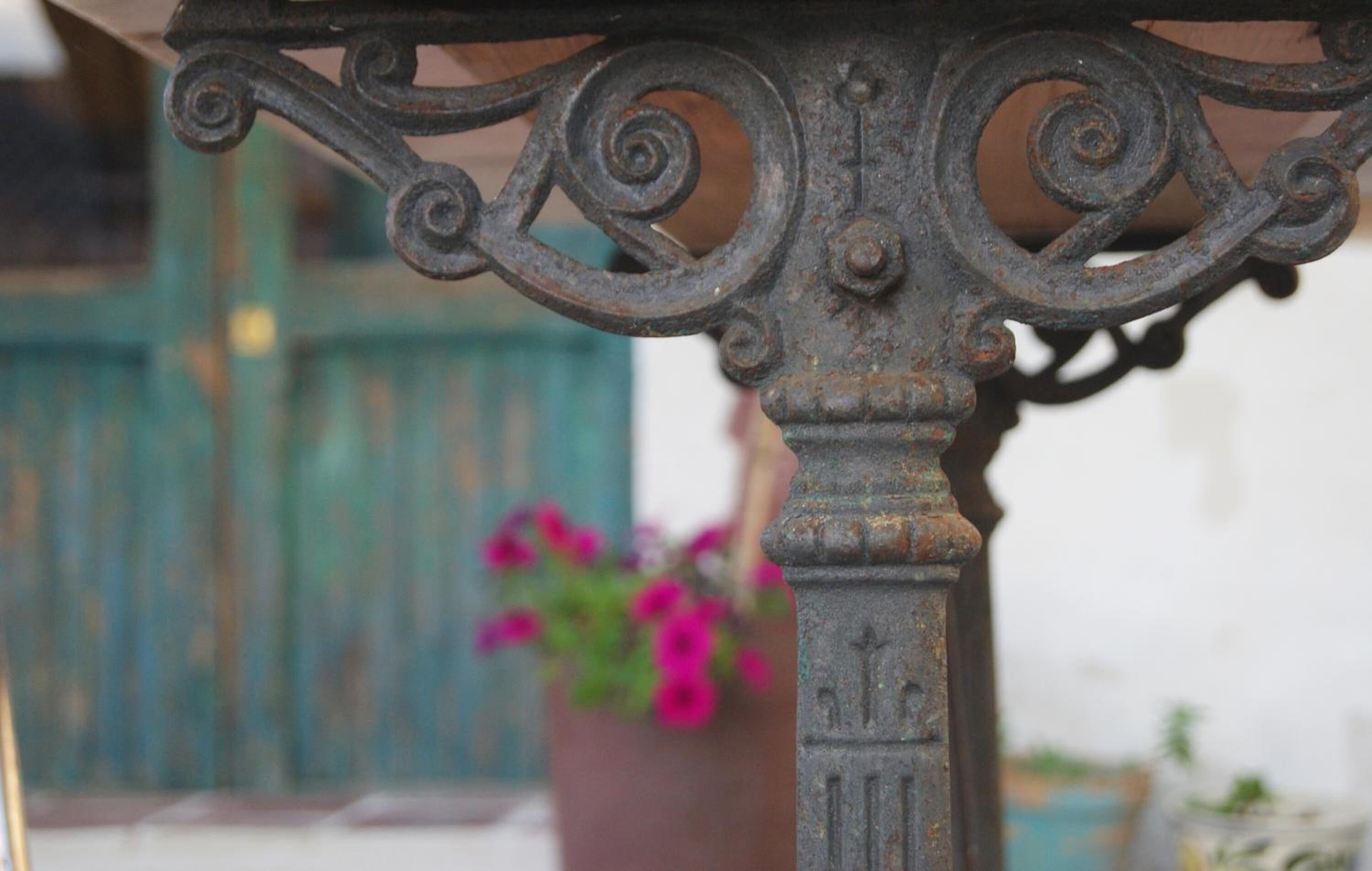 Un gros plan d’une table en métal patinée par les intempéries, ornée de motifs décoratifs complexes, avec une plante en pot aux fleurs roses, et un arrière-plan flou où l’on aperçoit une porte verte.