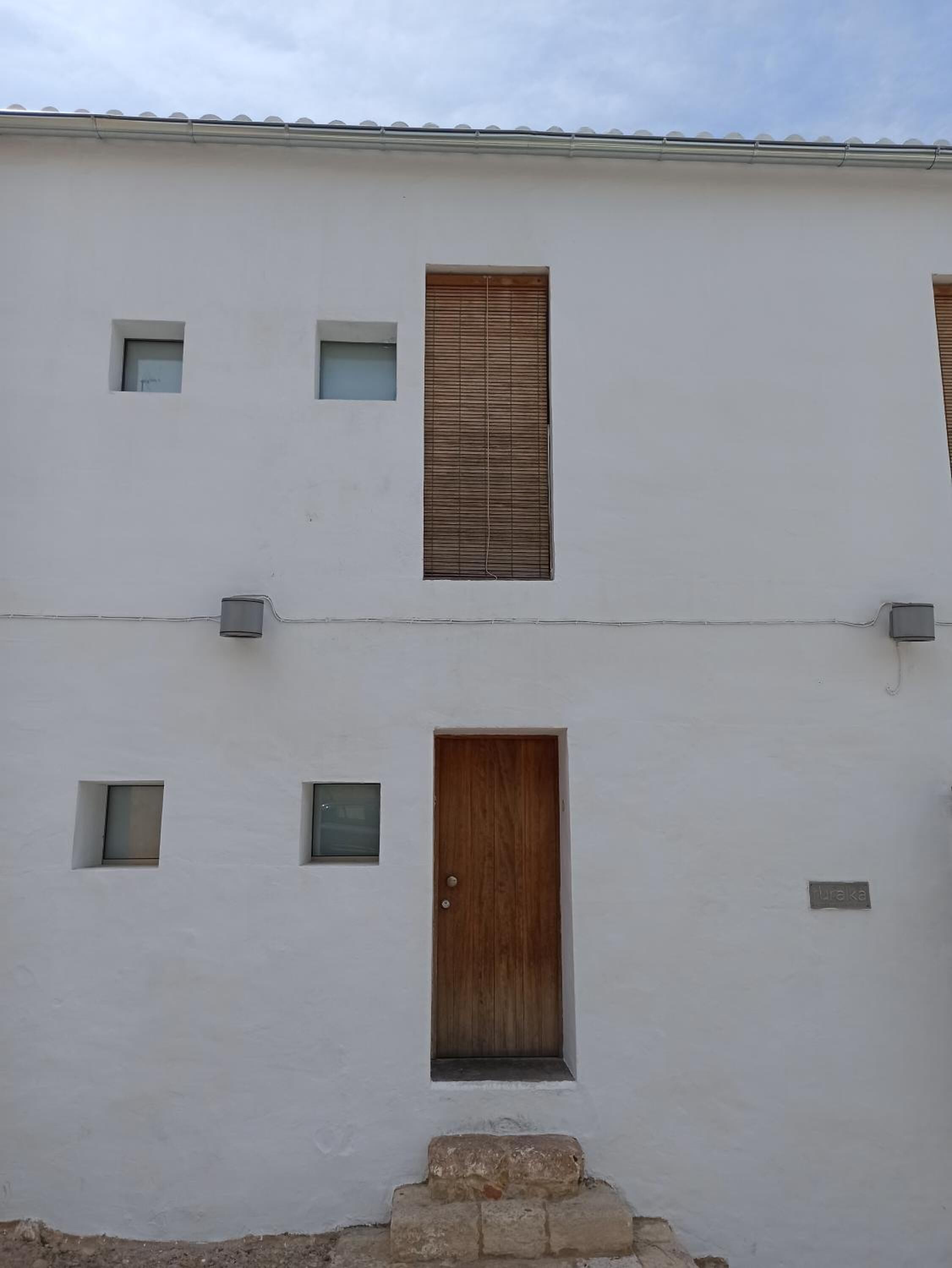 A white building with a central wooden door, two small windows above it, and a small window to the left.