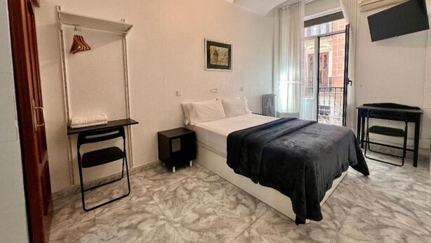 A bedroom with a bed, a small table, a hanging rack, and a window with a view.
