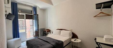 A hotel room with a bed, a small table, a chair, a TV, and a desk with a hanging coat rack.