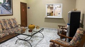 A hotel room with a sofa, armchair, and a glass coffee table. There is a painting on the wall, a door, and a window with a view.