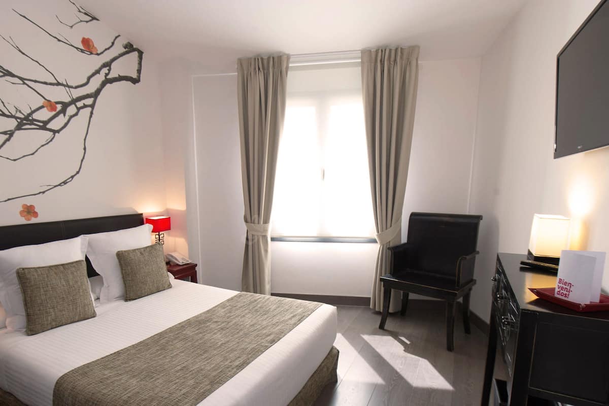 Une chambre d’hôtel avec un lit, un bureau, une chaise, une télévision et une fenêtre avec des rideaux.