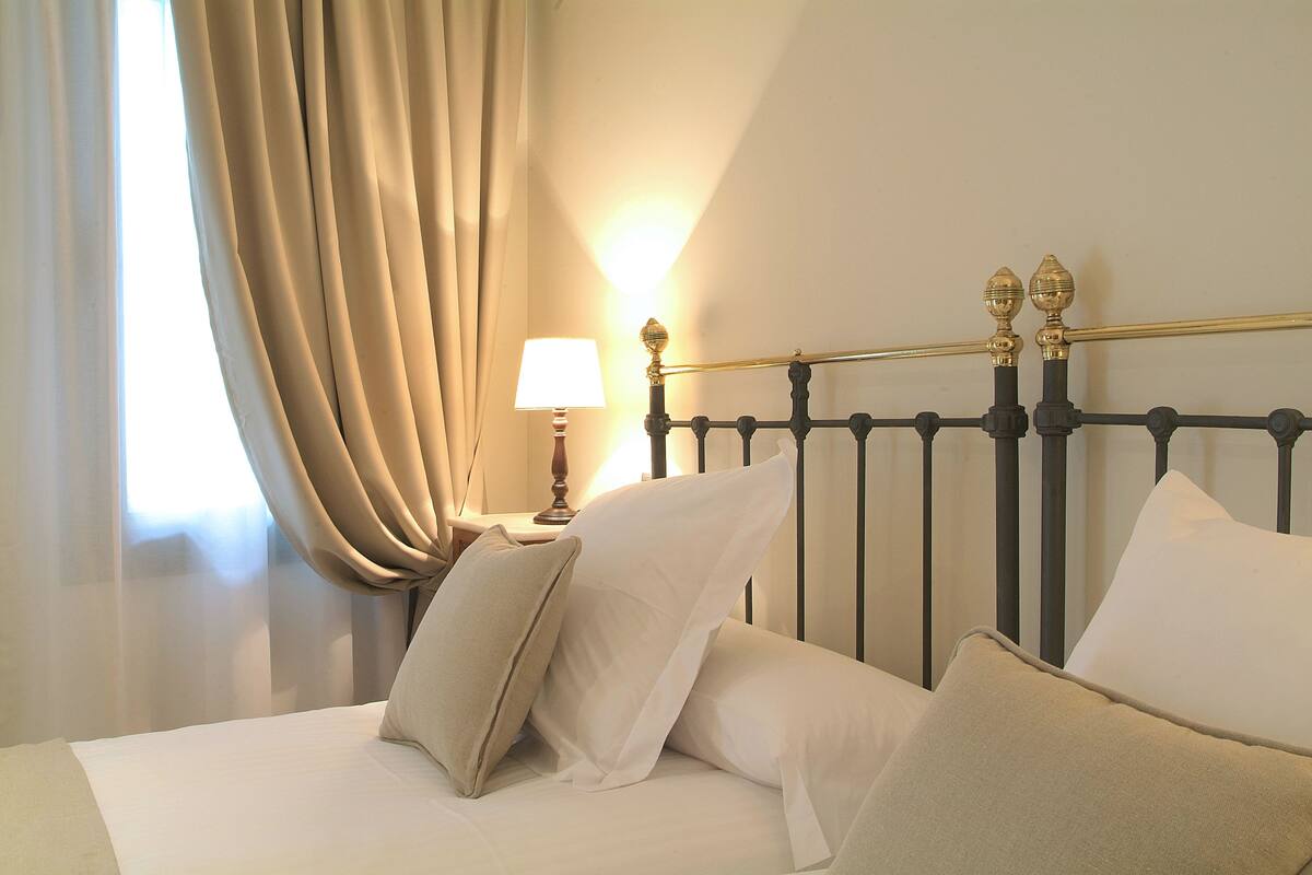 Un lit bien fait, avec du linge de lit blanc et des oreillers beige, une lampe de chevet et une fenêtre avec des rideaux.