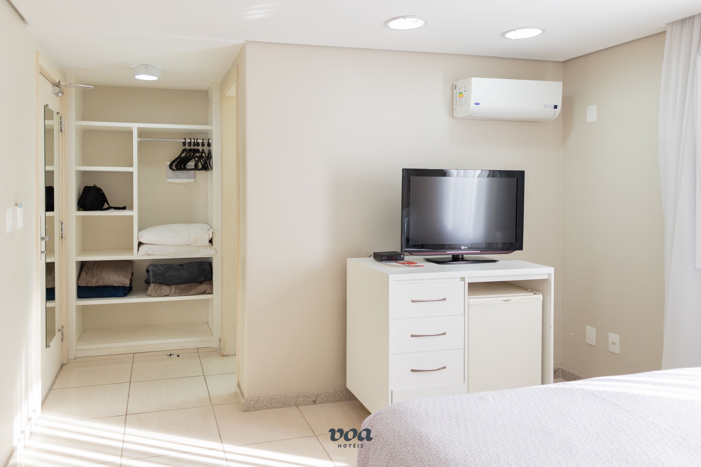 A hotel room with a bed, a TV on a white cabinet, a closet with hanging clothes, and a window with curtains.
