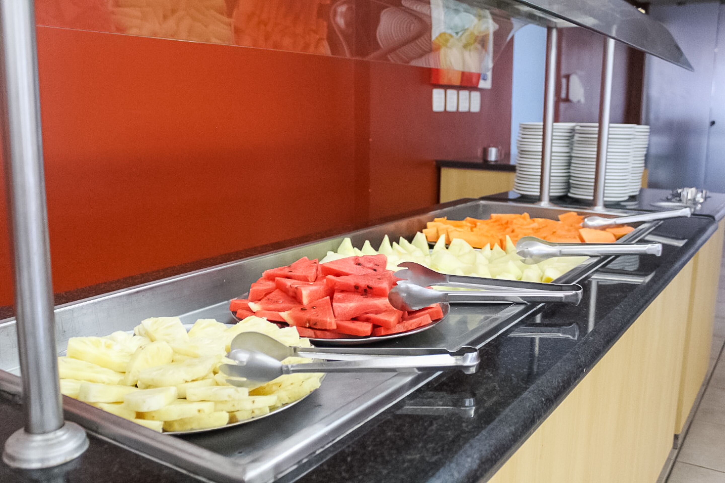 Een buffet met diverse soorten fruit en groenten, waaronder ananas, watermeloen en wortels, gepresenteerd op metalen dienbladen met bestek.
