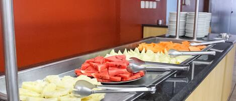 Een buffet met diverse soorten fruit en groenten, waaronder ananas, watermeloen en wortels, gepresenteerd op metalen dienbladen met bestek.