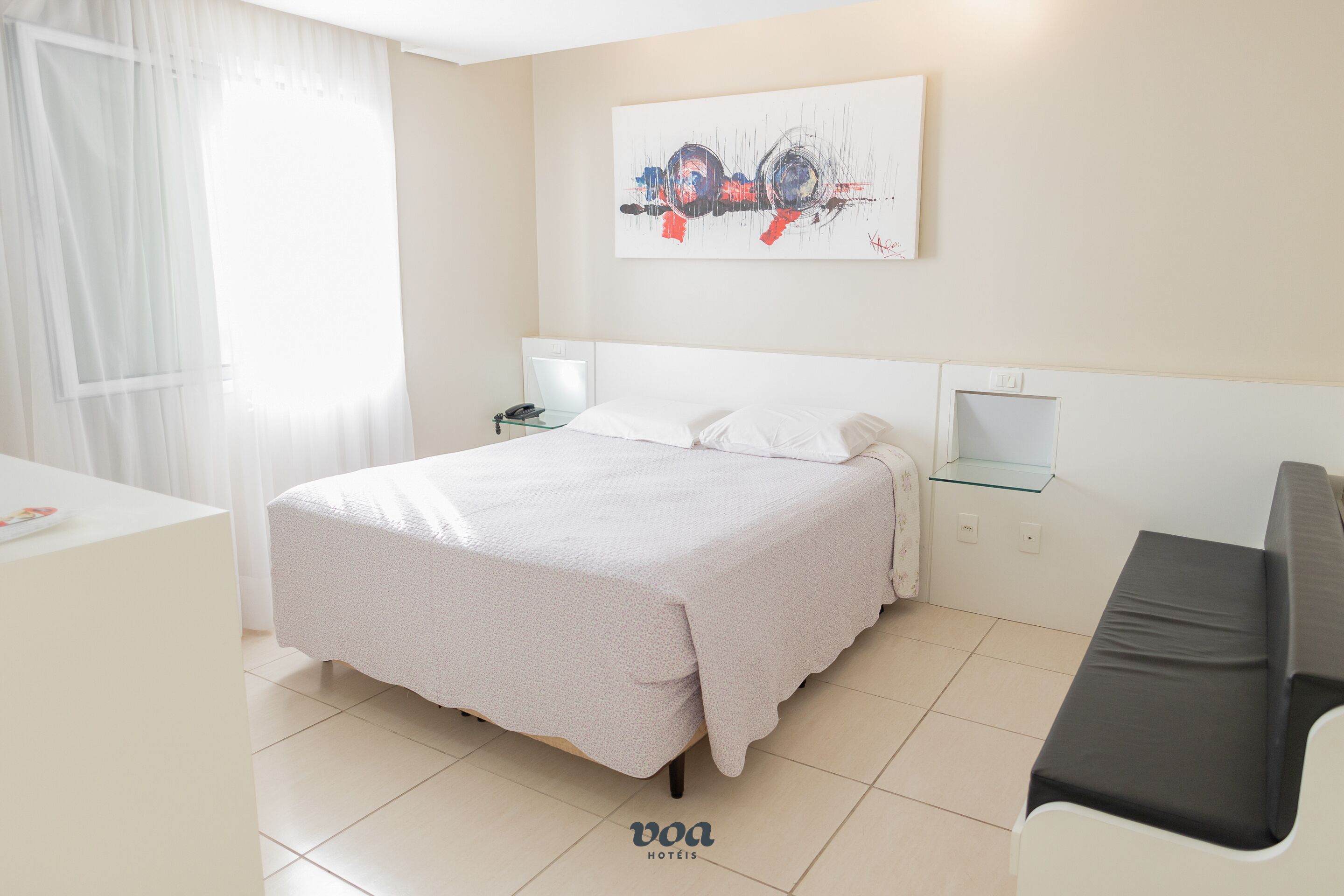 A hotel room with a bed, a desk, a chair, a nightstand, and a wall-mounted lamp.