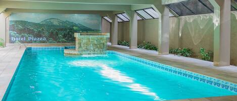 An indoor swimming pool with clear blue water, a stone fountain, and a mural of a mountainous landscape.