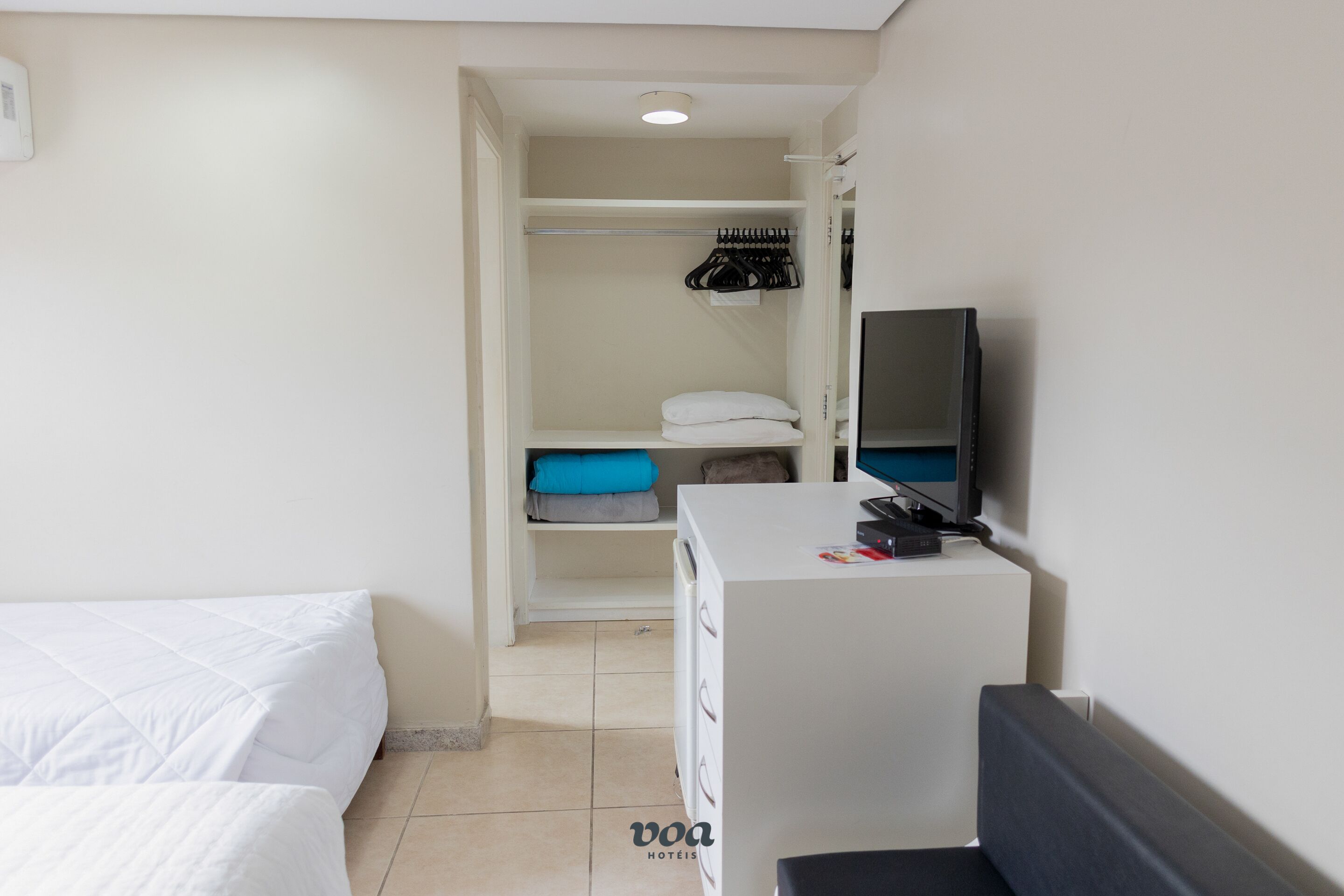 Quarto de hotel com cama, escrivaninha com computador, televisão e armário com roupas penduradas.